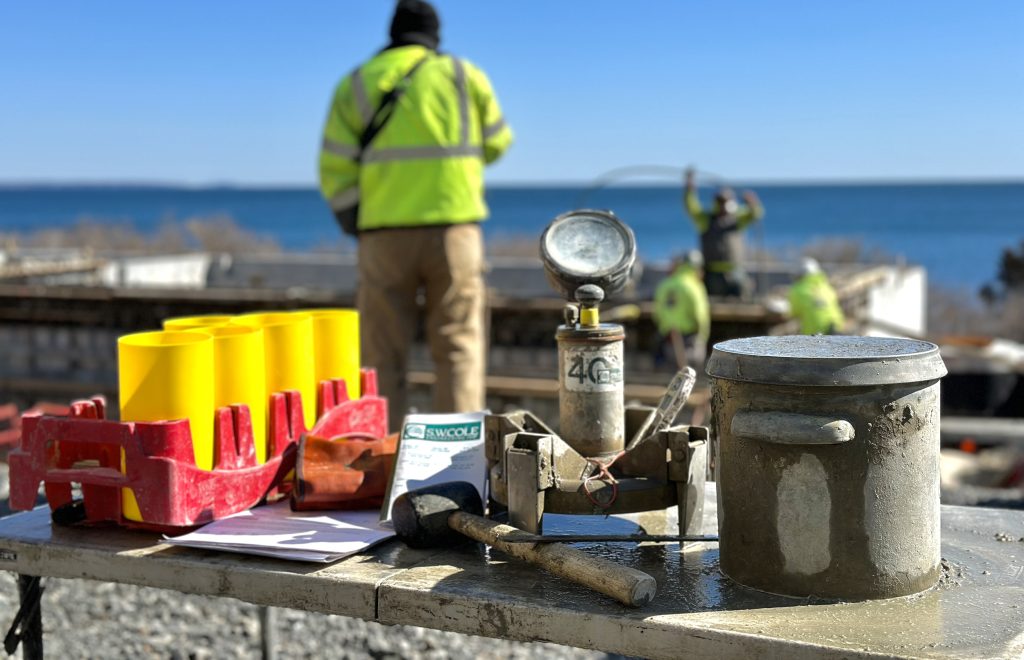 Concrete testing tools laid out on a table overlooking a construction site with people at work and the ocean beyond.