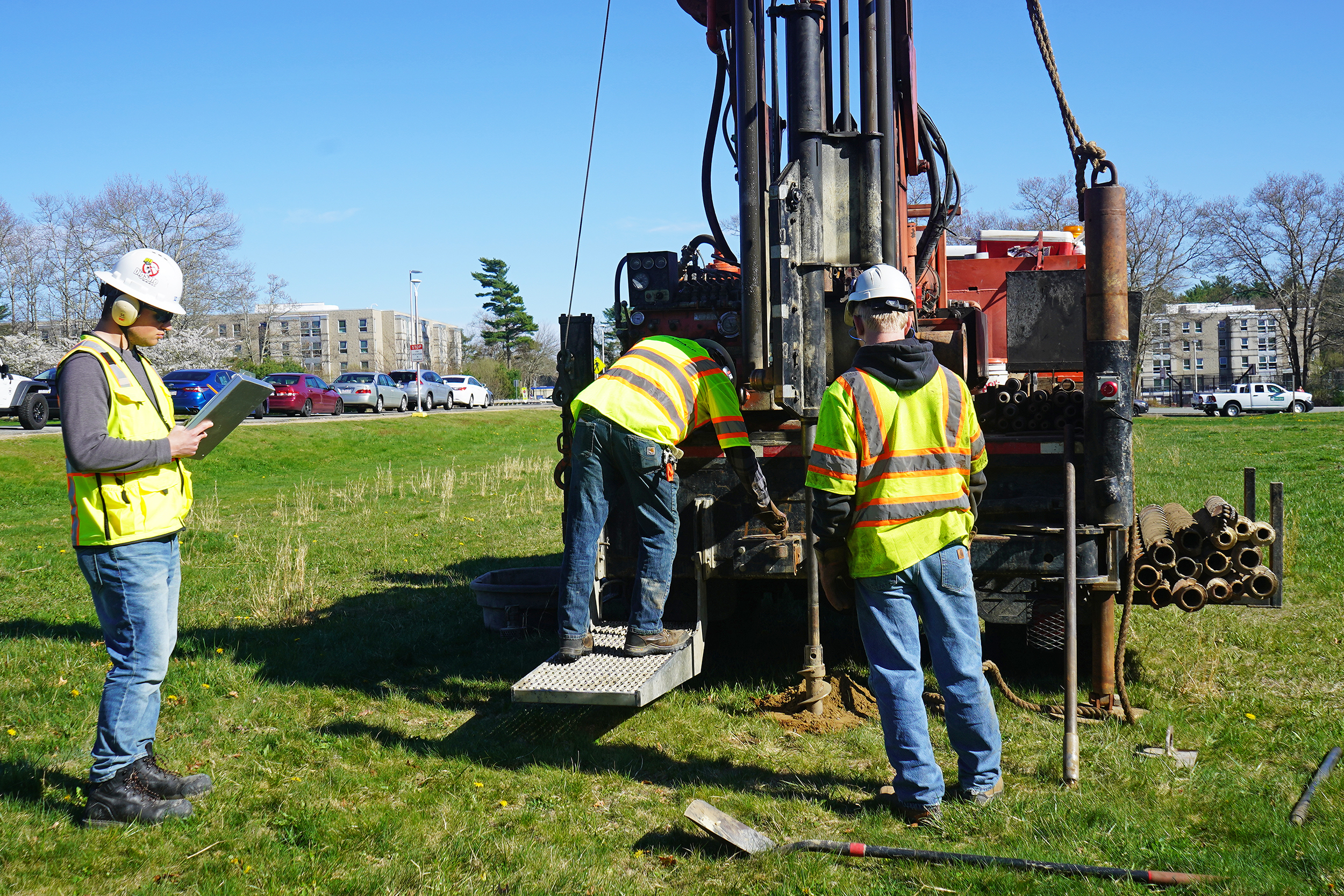 A geotechnical engineer writes on a clipboard while observing a drill rig and crew.