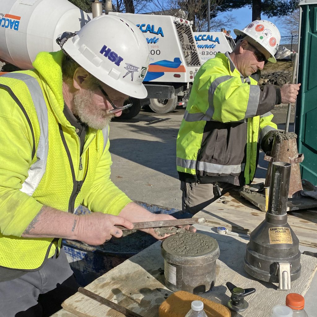 Two men wearing high visibility clothing and hardhats performing tests on concrete