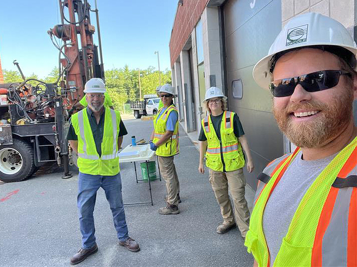 A group of employees facing camera and smiling at geotechnical engineering project with drill rig in the background