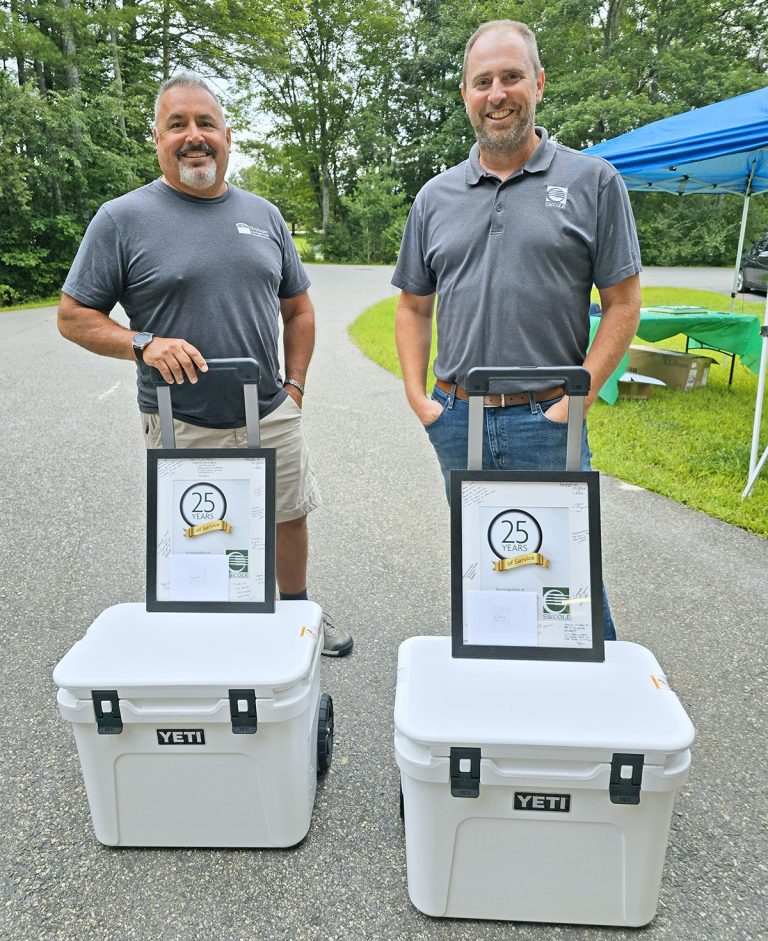 Two men stand outdoors on a paved surface, each smiling and posing with a framed certificate and a white YETI cooler. The certificates display a “25 Years” emblem, celebrating their service anniversaries. A blue canopy is visible in the background, with trees lining the scene.