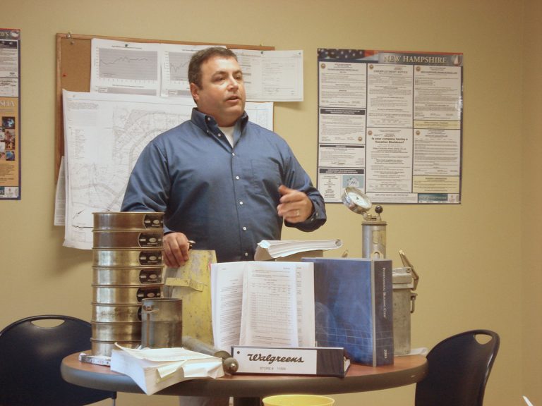 A man stands in a workspace, speaking and gesturing with one hand. He is surrounded by engineering materials, including a stack of metal sieves, and open reference books on a round table. The background features maps and technical posters pinned to a bulletin board.