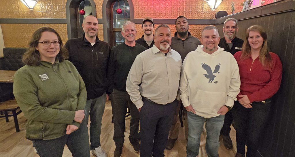A group of nine people stand together inside a warmly lit restaurant, smiling for the camera. The setting features wood floors and festive holiday decorations.