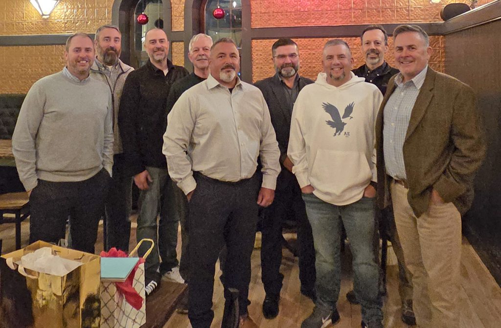 Nine people pose together in a restaurant, with a gift bag visible in the foreground. The group is dressed in casual and business attire, and the background includes arched windows and holiday ornaments.