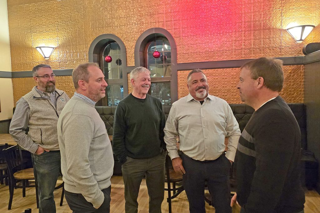 Five people stand in a semi-circle, engaged in conversation and smiling. The restaurant setting features wood floors and cozy lighting.
