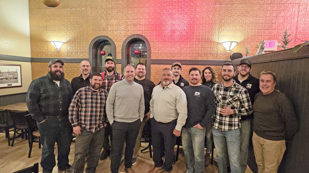 A larger group of thirteen people gather for a group photo in a restaurant. Most are dressed casually, and the atmosphere is cheerful with warm lighting and festive decor.
