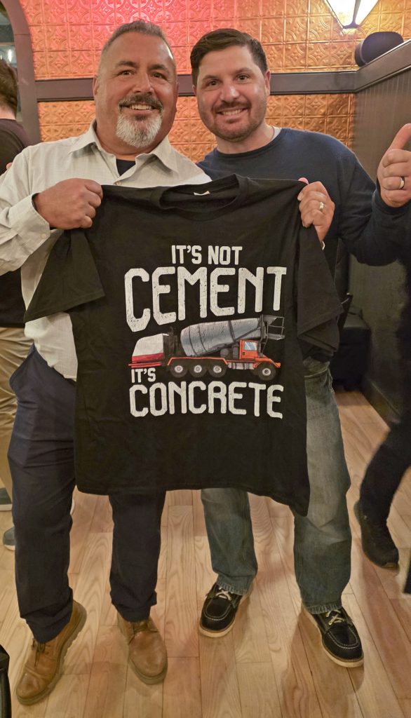 Two men pose together, holding up a black t-shirt that says “It’s not cement, it’s concrete.” Both are smiling, and one gives a thumbs-up gesture.
