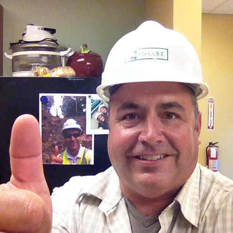 A man wearing a white hard hat and a light plaid shirt smiles and gives a thumbs-up gesture in an office setting. Behind him, a refrigerator displays a printed photo of a man at a construction site, also giving a thumbs-up. The background includes a fire extinguisher and kitchen appliances.