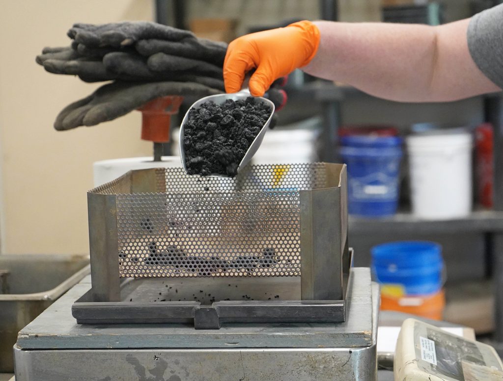 A construction materials testing technician is emptying a scoop of asphalt onto a scale.