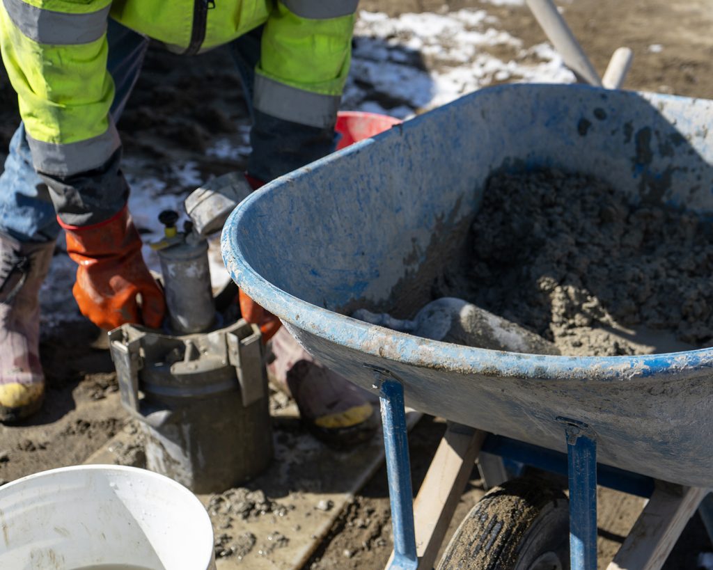 A wheel barrow with fresh concrete is in the foreground and a construction materials technician is performing an air content test in the background.