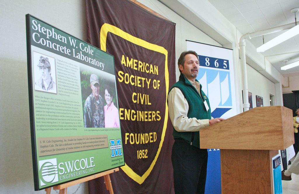 Paul Kohler speaking at an event with displays behind him representing the University of Maine, the American Society of Civil Engineers, and the Stephen W. Cole Concrete Laboratory.